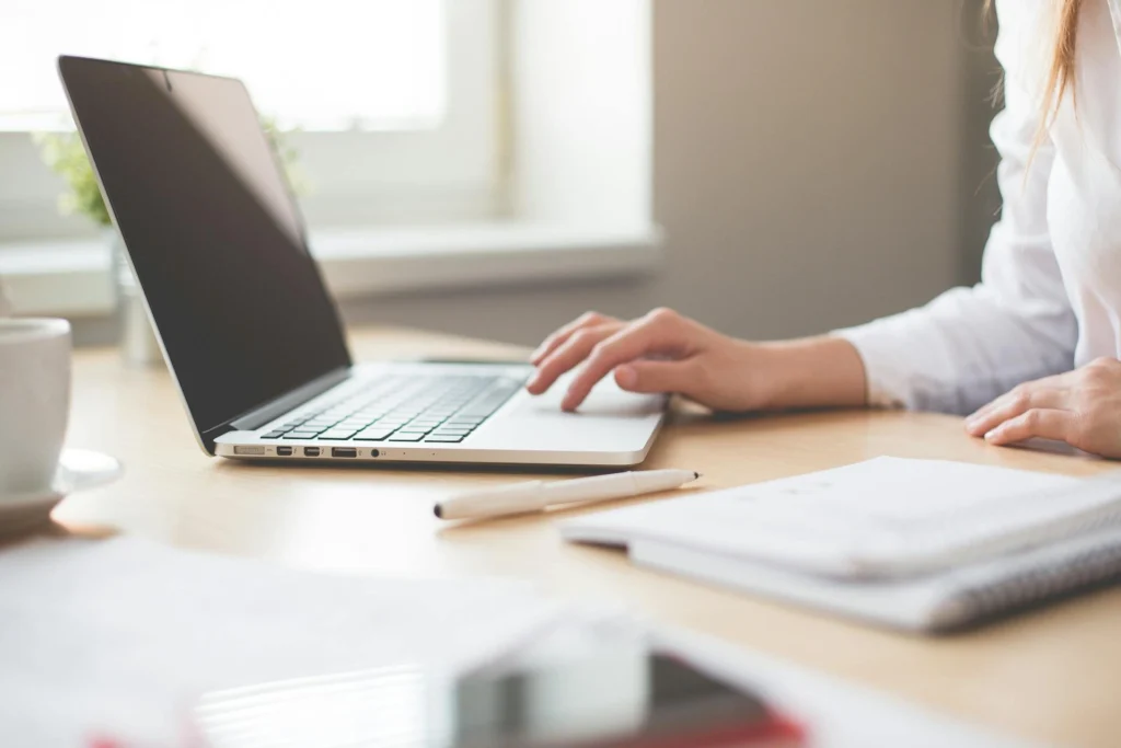 In the image you can see the hand of a woman working on a laptop. Next to it you can see a pen, a notepad and a cup.