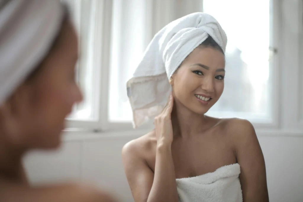 In the picture a girl looks at herself in the mirror and smiles. She has a white towel over her hair and is covered with another towel.